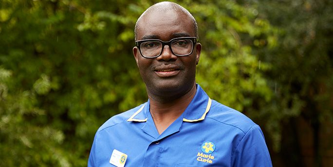 Marie Curie Nurse Isaac smiles as he poses for a photograph outside near trees. He's wearing a blue Marie Curie uniform and black glasses.